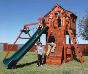 Kids playing in a redwood fort with a green slide, swings, a climbing wall and a small house on top.