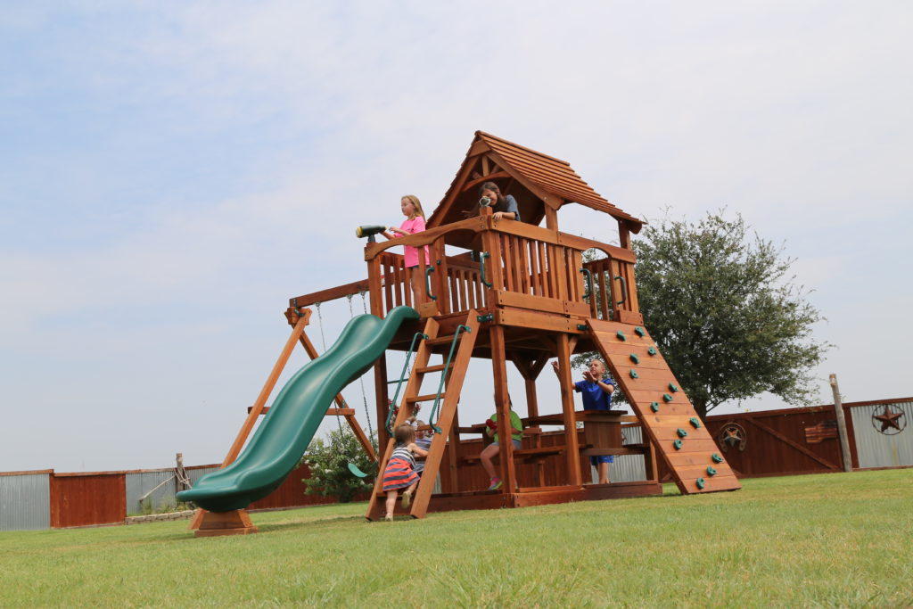 Kids playing in a redwood fort with a green slide, swings, a climbing wall and a small house on top, electronics forgotten only adventure play ahead.