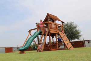 Kids playing in a redwood fort with a green slide, swings, a climbing wall and a small house on top.