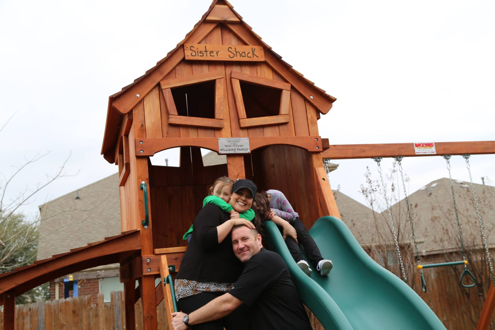 A picture of one of Backyard Fun Factory's Amazing Love Giveaway redwood playsets - featuring a fort, slides, swings, and, of course, the happy family.