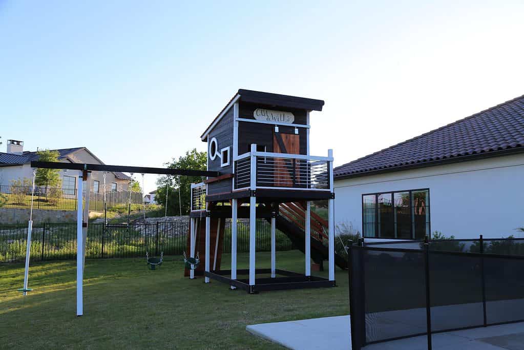 ebony stain and white paint trim modern design playset with circle and square shaped windows, front and back deck, upper cabin with sliding door, ramp, swings and climber.