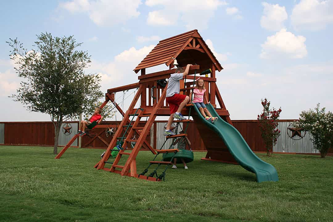 Haslet Texas, 5' deck height, fort concho swing set with rope ladder,Kids playing in a redwood fort with a green slide, swings, a climbing wall and a small house on top.
