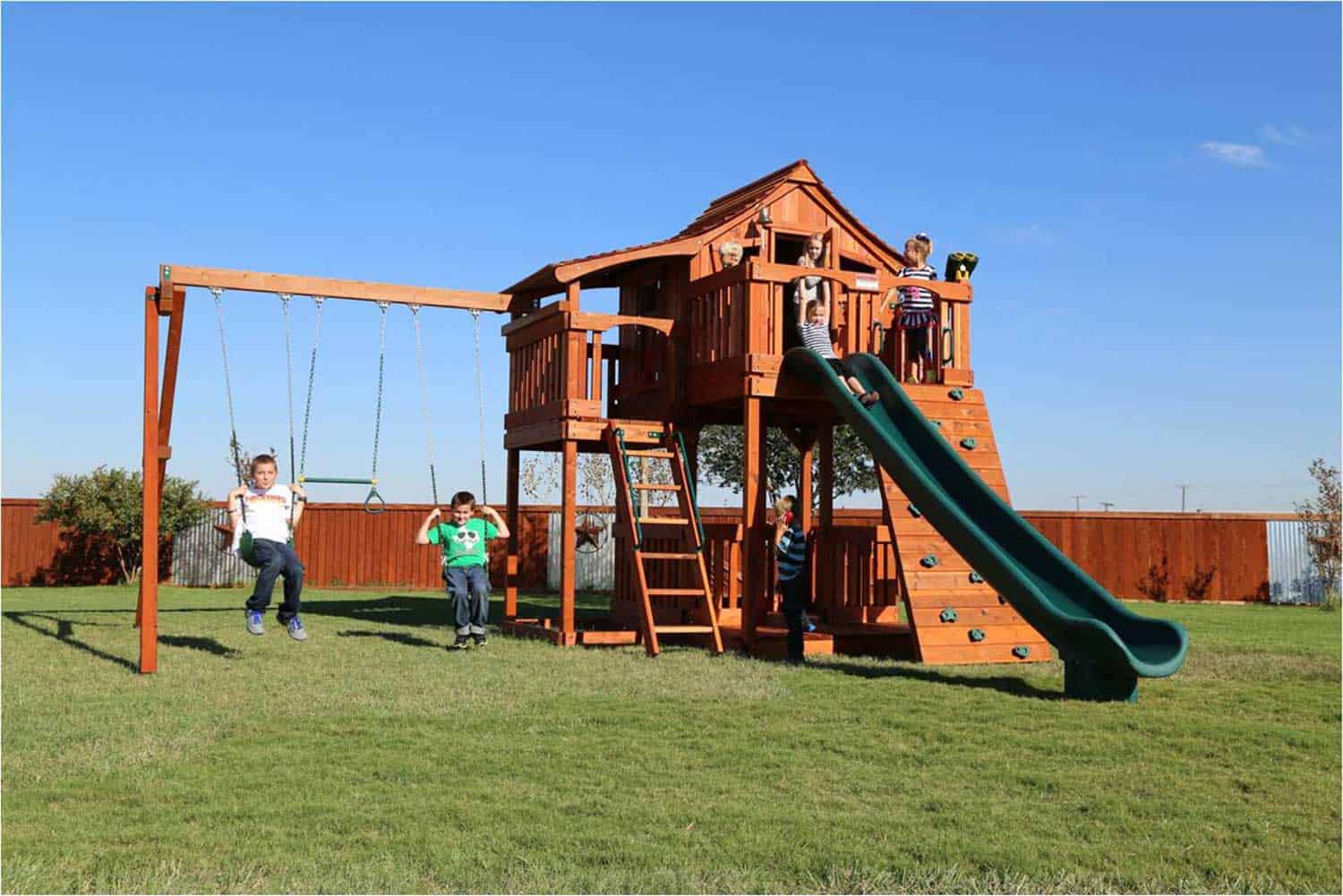 fort stockton swing set shown with 4 foot half shack, upper cabin, patio, fun deck, kids swinging and sliding, rock wall