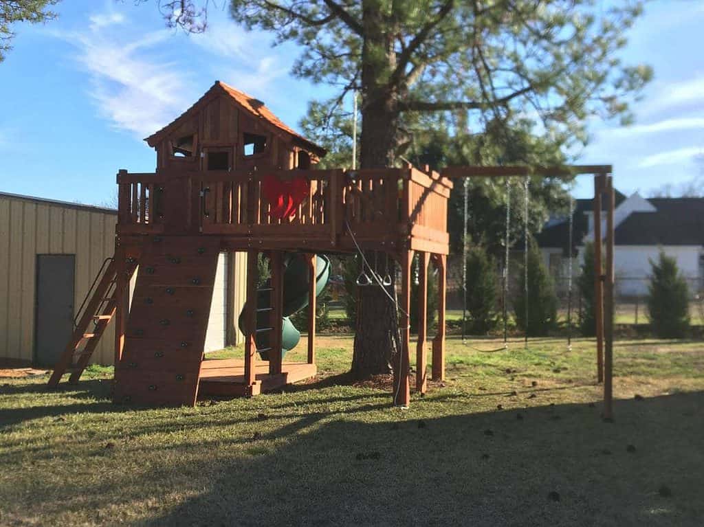 Christmas gift of redwood playset with big red bow on front. playset has cabin, rock wall, tree decks, swings and spiral slide.
