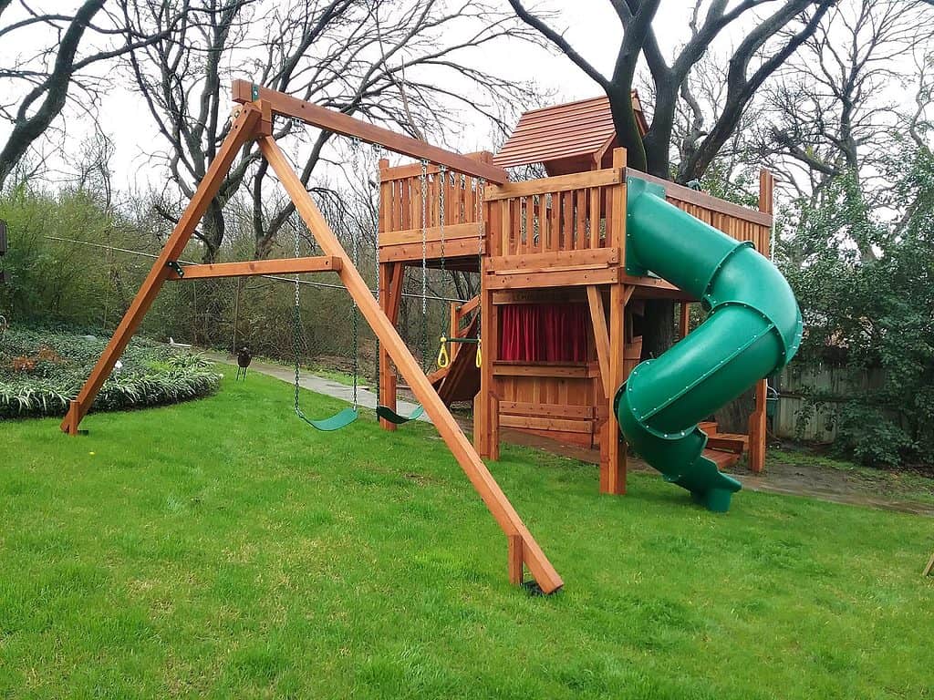fort with tree deck installed on slope, wooden playset customized around a backyard tree with swing set accessories, cabin and slides