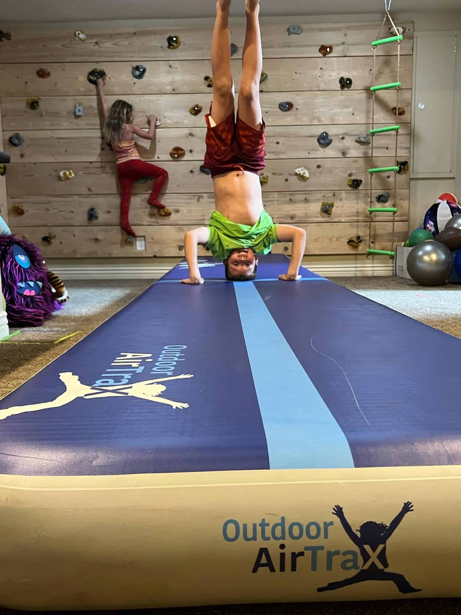Arlington Texas, Young boy performing gymnastic head stand on air trax in an indoor room where young girl is climbing rock wall in background.