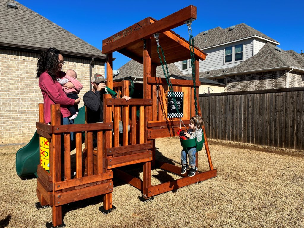 family playing on toddler trainer redwood playset with mom holding baby and toddler swinging in the toddler swing