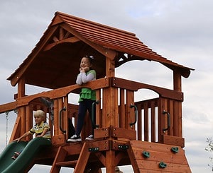addition swing set component Open slat fort shown with half slatted walls and wooden roof. Little girl standing on deck with little boy sitting on slide