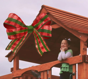 Girl smiles, hands on her chin, looking out from the top of a redwood covered deck with a large Christmas bow on top.
