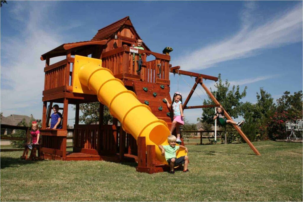 Joyful children enjoying themselves on a large Backyard Fun Factory playset, featuring a bright yellow enclosed slide, a fort, a lemonade stand, swings, and a climbing wall.