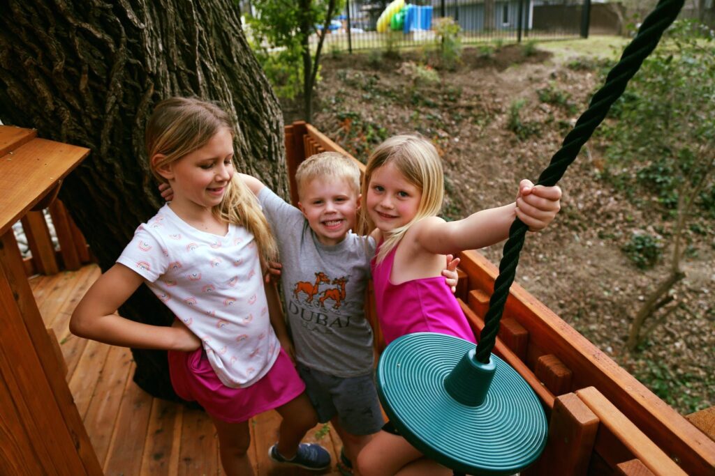 Two sisters pause their play for the camera, joined by their baby brother who smiles, as they laugh and enjoy their time on the redwood Backyard Fun Factory playset."