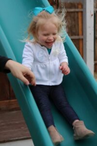 The science of speed is found in STEM outdoor playing on a slide. A young blonde child beams with joy as she glides down a vibrant green Rocket slide, while her father's steady hand offers gentle reassurance nearby.