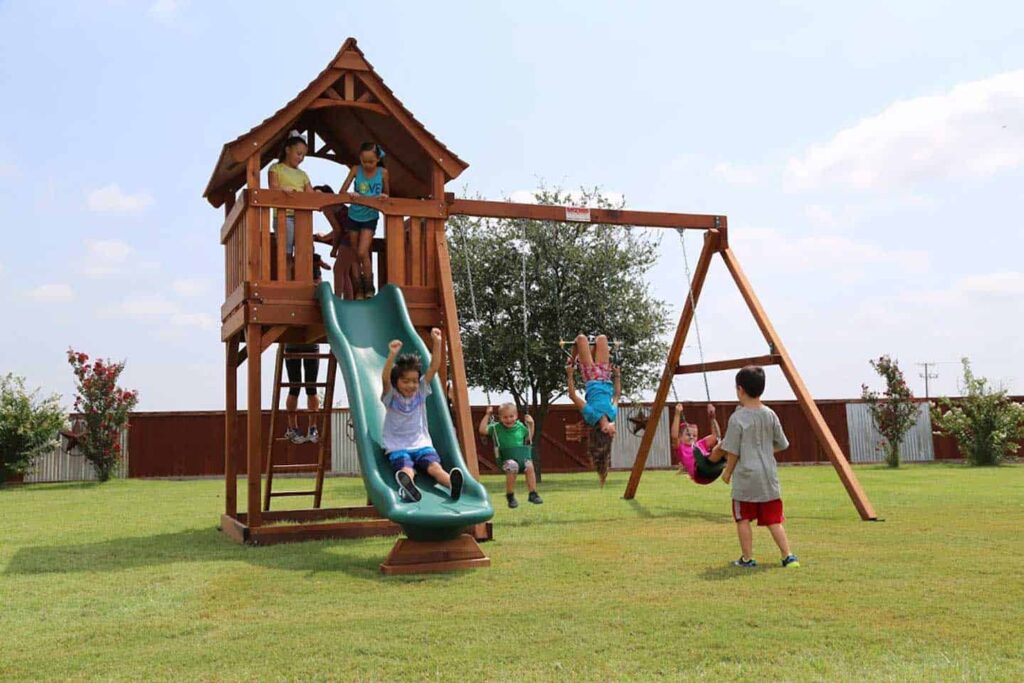 Joyful children enjoying themselves on a Backyard Fun Factory playset, featuring a green rocket slide, a covered deck, and a swing bar with two belt swings and a trapeze bar.