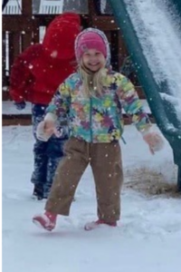A close-up of a young girl all bundled up smiling brightly as she plays in front of her Backyard Fun Factory playset in the snow.