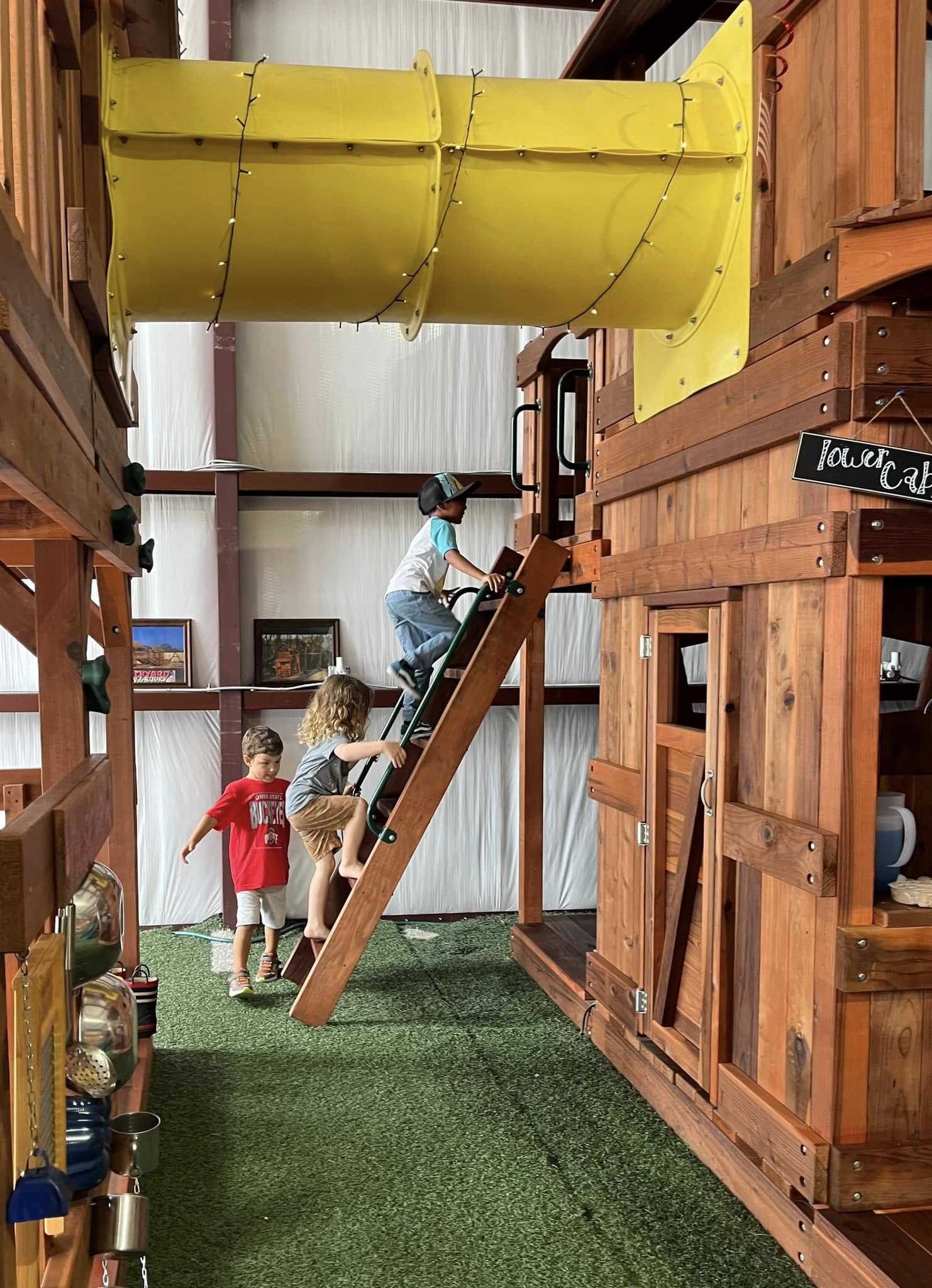 Three young boys play on a wooden play structure at an indoor playground. Two boys climb a wooden ladder toward a platform with a yellow tube slide, while another boy walks nearby on artificial grass. A sign reading "Tower Café" hangs on the structure, and play kitchen items are visible in the background, creating a fun and interactive environment.