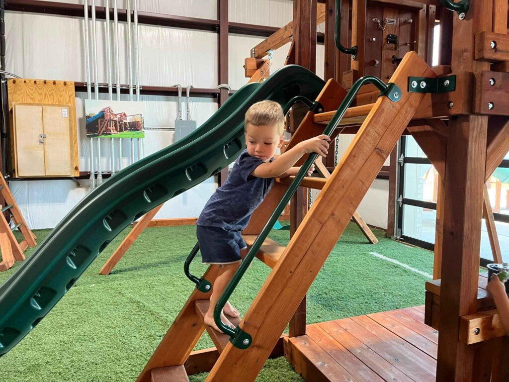 Young boy climbing wooden ladder on Fort Davis playset at Backyard Fun Factory Frisco Swing Set Showroom, featuring a custom redwood playset with a green slide and durable backyard play equipment for kids.