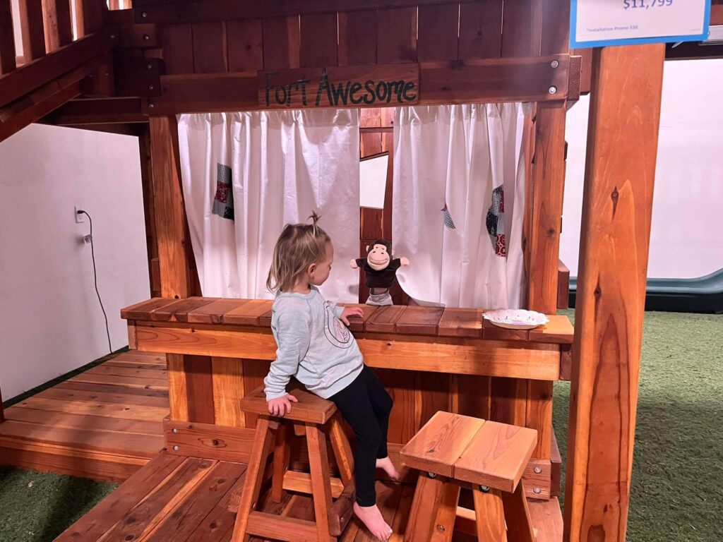 Young girl watching a puppet show at the Fort Awesome wooden playset in Backyard Fun Factory Frisco Swing Set Showroom, featuring custom redwood playsets with interactive play areas for kids.