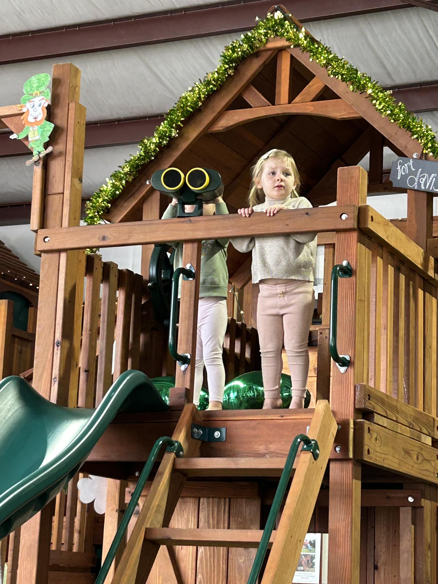 Two young girls play on a wooden playset named "Fort Davis" at an indoor playground. One girl looks out from the platform while the other peers through binoculars. The play structure is decorated with green garland and features a green slide. A leprechaun decoration and a sign reading "Fort Davis" are visible, creating a festive atmosphere.