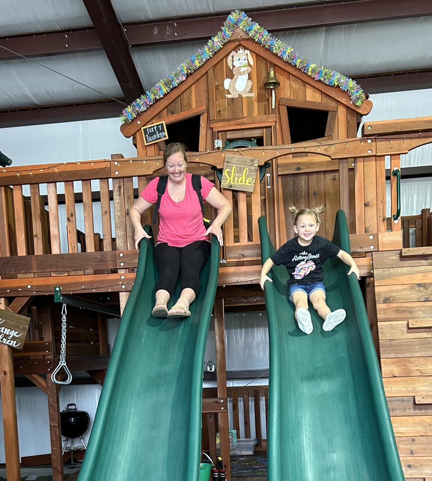 A woman and a young girl race down parallel green slides on a wooden play structure at an indoor playground. Both are smiling and enjoying the moment. The playset is decorated with colorful garland, a "Slide" sign, and a small house structure labeled "Fort Tucker," creating a fun and family-friendly atmosphere.