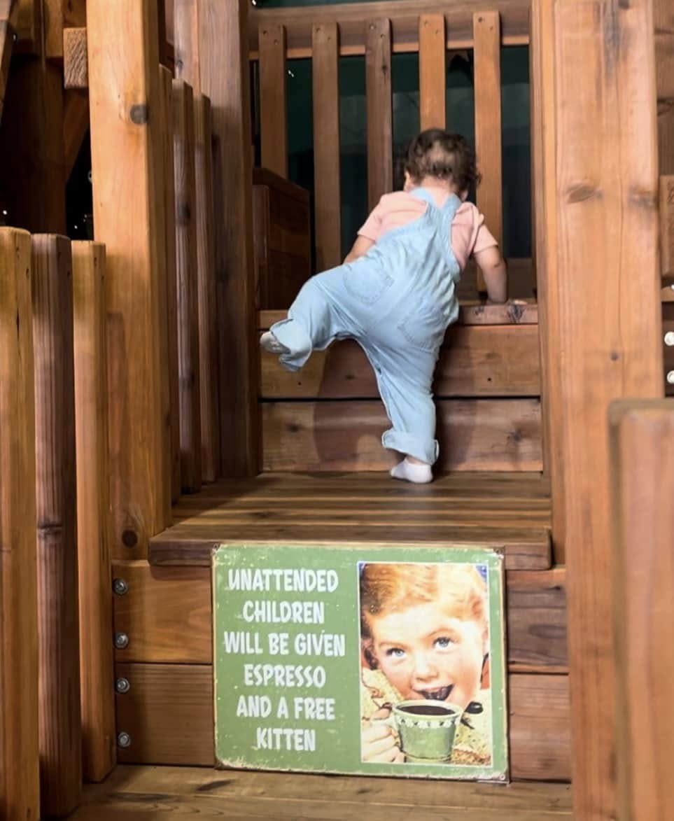 A toddler climbs wooden steps on a play structure at an indoor playground. A humorous sign at the base of the stairs reads, "Unattended children will be given espresso and a free kitten," accompanied by a vintage-style image of a child holding a cup. The wooden playset creates a warm and playful atmosphere.