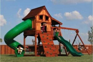 Excited children play on a beautiful redwood playset with a sturdy swing beam a green Rave and Spiral slides, front Climbing wall that goes up into a large upper fort with side deck and a lower deck.