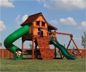 Excited children play on a beautiful redwood playset with a sturdy swing beam a green Rave and Spiral slides, front Climbing wall that goes up into a large upper fort with side deck and a lower deck.