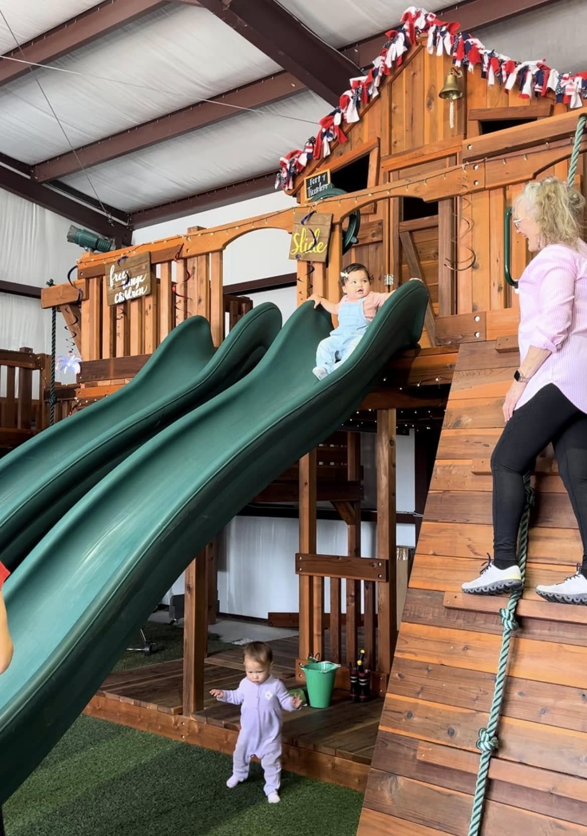 Two toddlers play on a wooden play structure at an indoor playground. One toddler slides down a green slide while another stands nearby on artificial grass. A woman climbs a wooden climbing rappel wall with a rope next to the slides. The playset is decorated with red, white, and blue ribbons and a sign reading "Slide," creating a fun and welcoming environment.