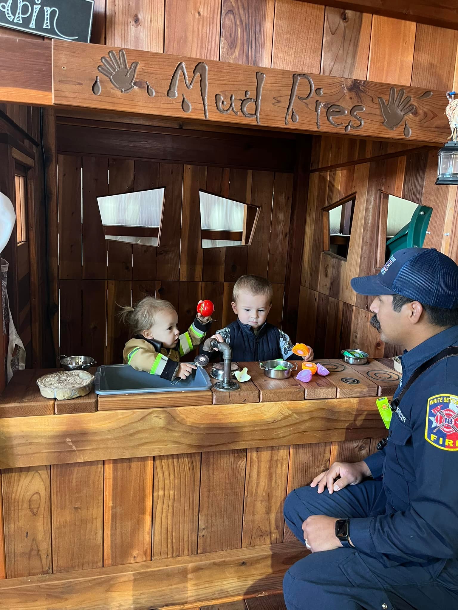 A firefighter interacts with two young children playing at a wooden mud kitchen playset labeled "Mud Pies" at an indoor playground. One child wears a firefighter costume and holds a toy vegetable, while the other plays with toy kitchen items. The firefighter, dressed in uniform, watches them with a smile, creating a playful and engaging atmosphere.