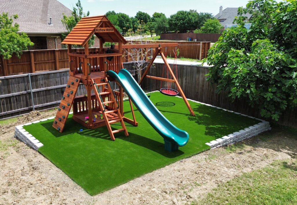 Backyard playground with wooden Fort Davis playset featuring a green slide, rock climbing wall, swing set, and artificial turf base, surrounded by a wooden fence and landscaping.