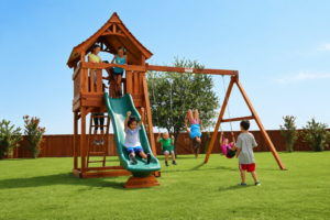 Joyful children enjoying themselves on a Backyard Fun Factory playset, featuring a green rocket slide, a covered deck, and a swing bar with two belt swings and a trapeze bar.